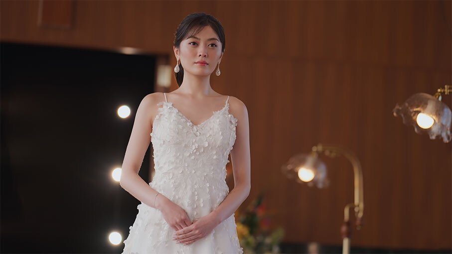 複数のライトに照らされた屋内に、白いドレスの女性が立つ。お腹のあたりで手を重ねている。