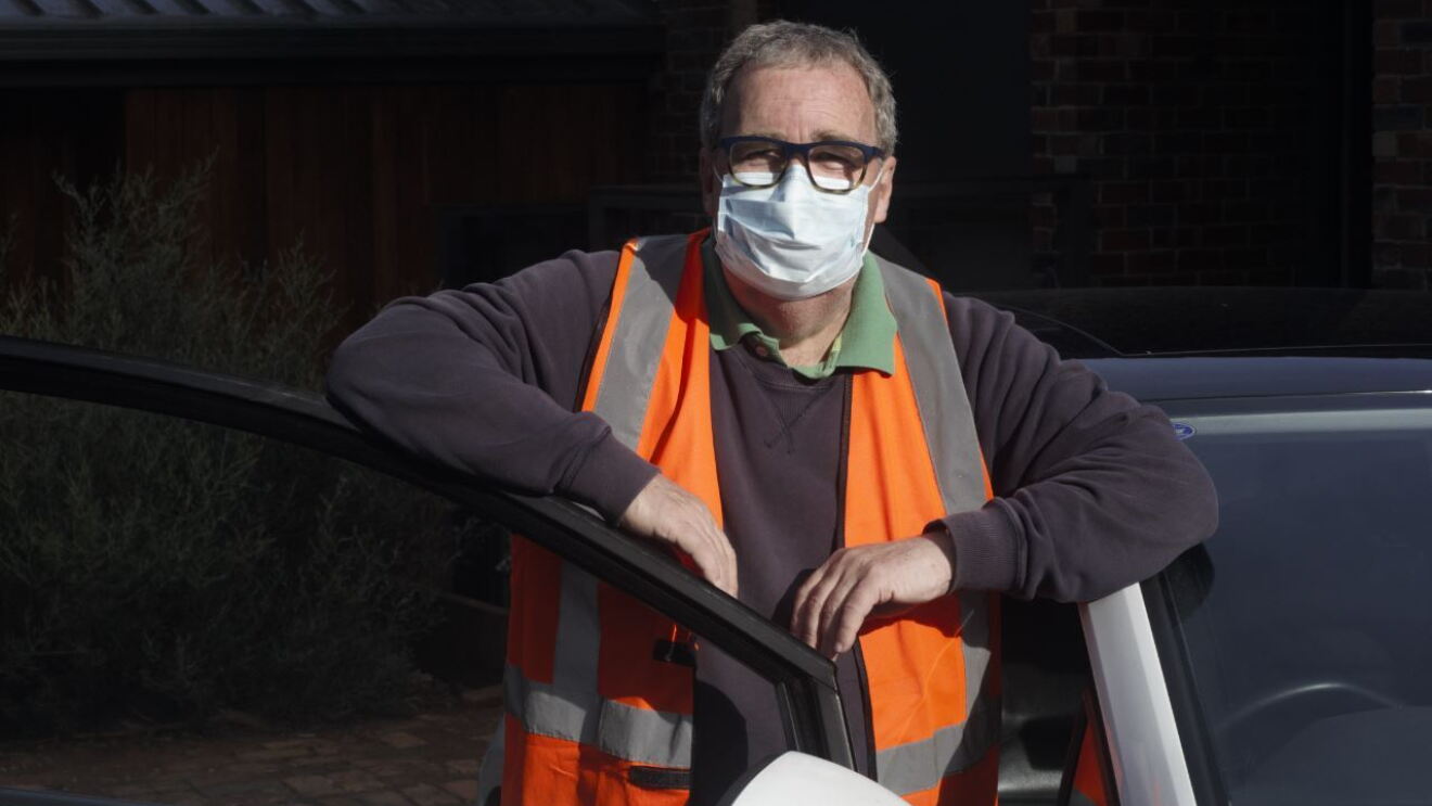 An image of a man next to his car smiling for a photo with a mask and an orange safety vest on. 