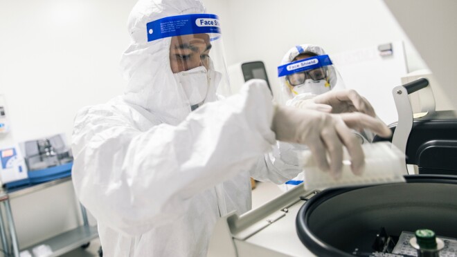 两人穿个人防护服。前景中的人把一个托盘放在一件实验室设备里。