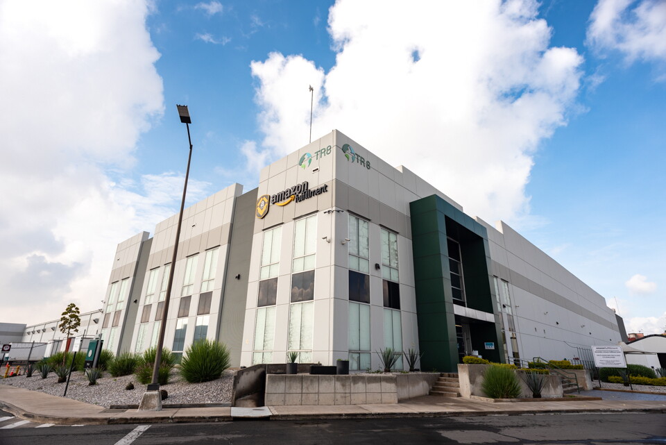 Centro logístico Mex1 en México. Foto exterior del edificio que tiene ventanas blancas, el logo de Amazon Fulfillment. El cielo está azul con nubes blancas. En la calle no hay coches y hay una acera con plantas.