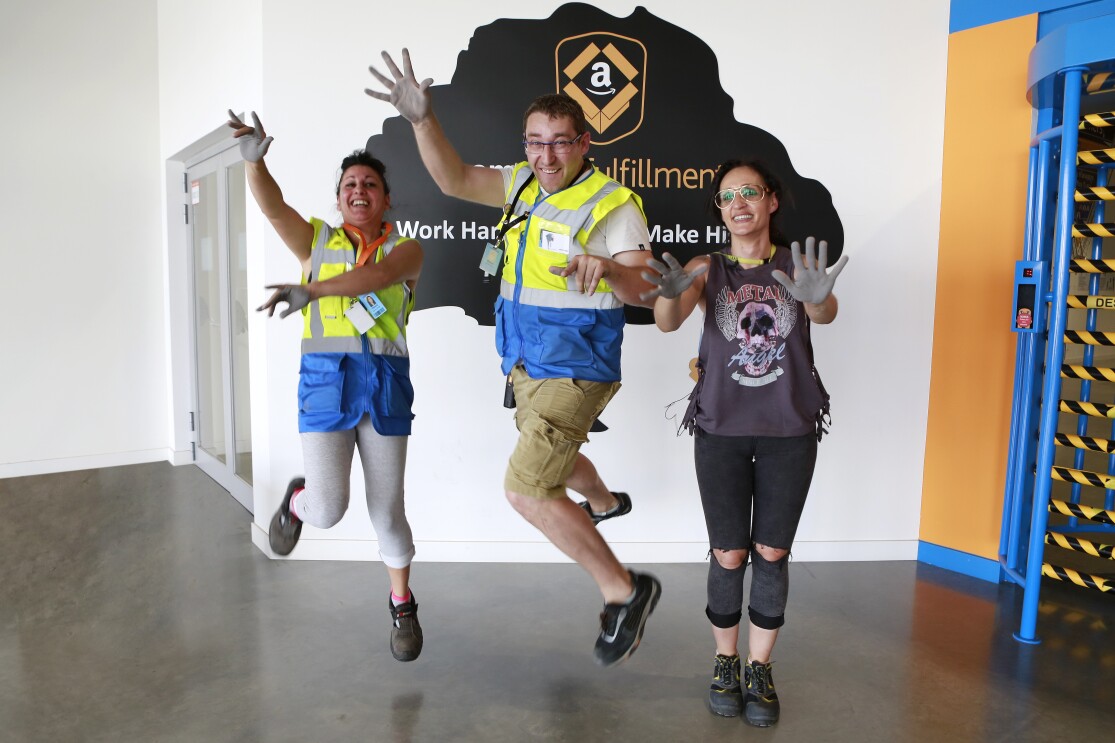 Patricia Alonso, Maribel Salcedo y Marius Dumitrescu aparecen en la entrada del centro logístico saltando y mostrando las palmas de sus manos pintadas de color gris. Están sonriendo mirando a cámara. Marius se enceuntra en medio.