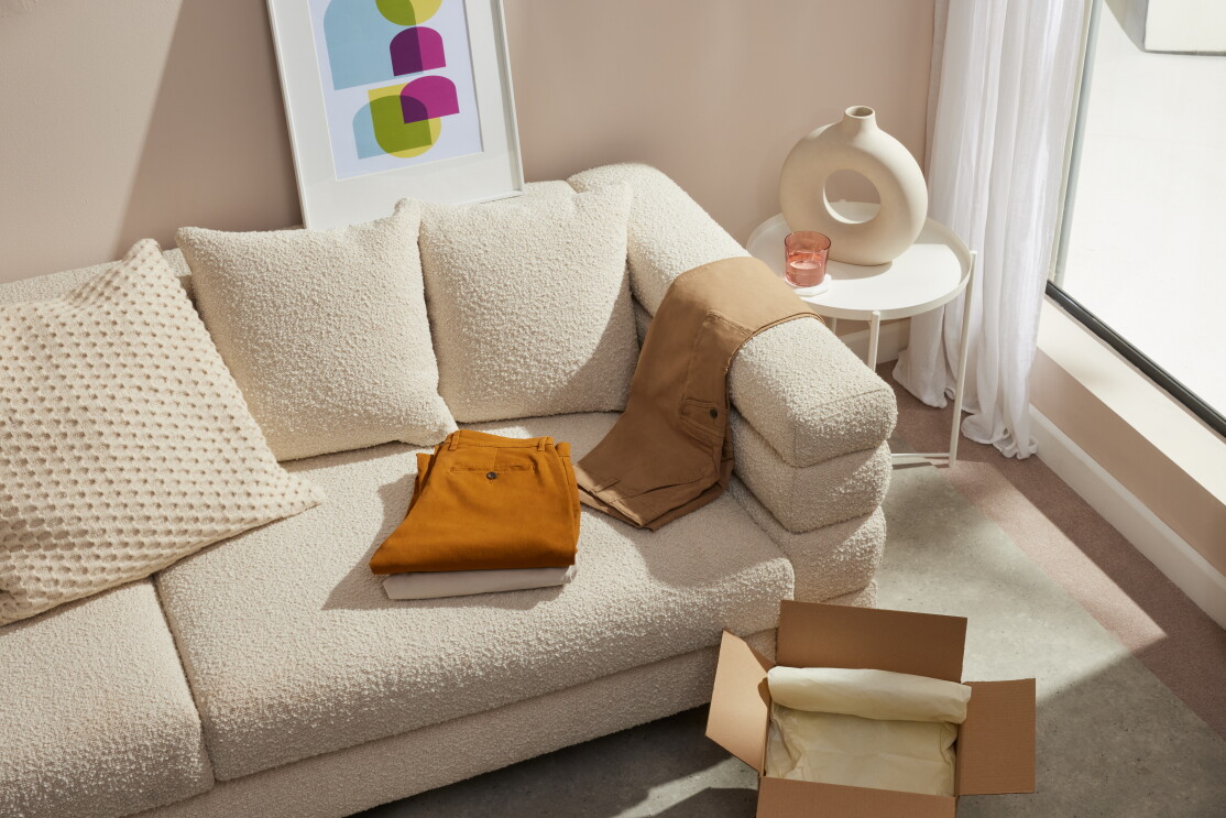 En el salón de una casa hay una sofá con un cojín de color beige,con tres cojines del mismo color. En el sofá hay un cuadro apoyado con unas figuras geométricas de color azul, morado y amarillo. Hay una ventana con una cortina de color blanco. Entre el sofá y la ventana hay una mesa redonda baja de color blanco, un vaso de cristal rosa transparente y un jarrón. En el suelo hay una caja marrón abierta con papel de envolver. Sobre el sofá dos pantalones plegados, uno beige y otro marrón. Apoyado en el brazo del sofá, otro pantalón de color marrón.