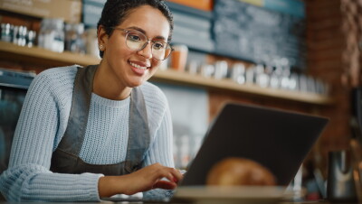 Uma mulher usando óculos está trabalhando em um computador. Ela está de suéter azul claro com um avental cinza