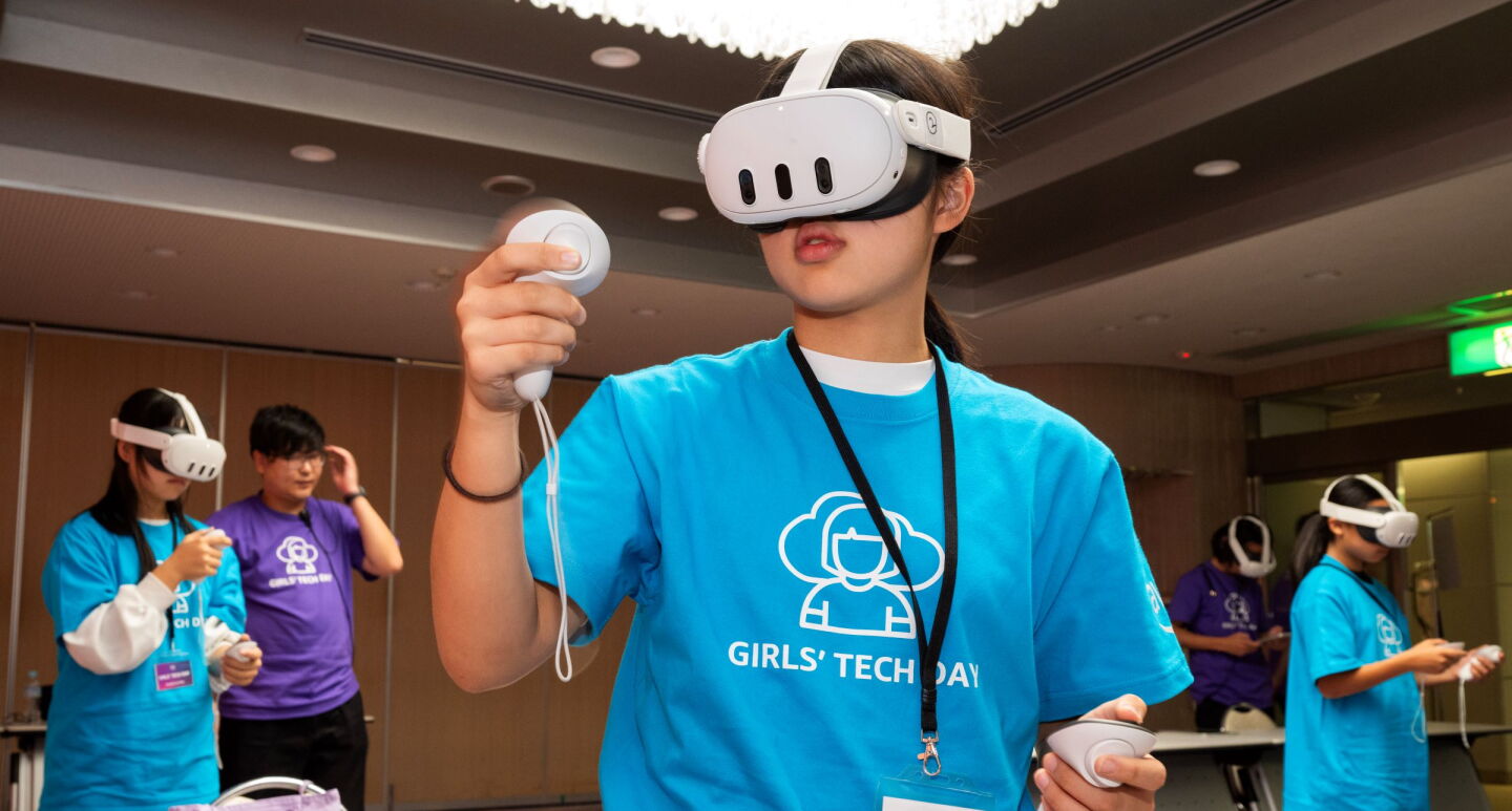 青いTシャツを着ている女性がVRヘッドセットをかけてコントローラーで動いているようだ。彼女の後ろには他の女性3人が同じことをしている。