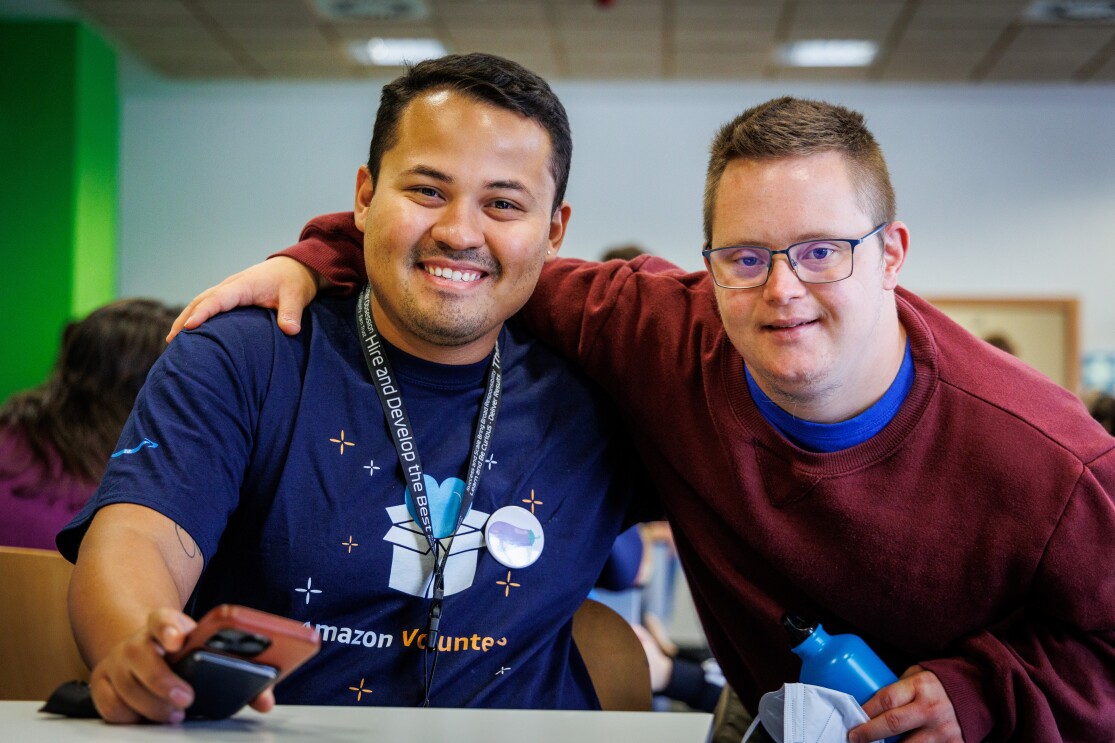 En las instalaciones de Down Madrid en Madrid aparecen 2 personas en primer plano. Un trabajador de Amazon y un participante de la fundación . El trabajador de Amazon van con la misma camiseta azul y el badge colgado y el de Down Madrid con deportivas con un jersey de color morado. La camiseta del trabajador de Amazon lleva un dibujo que es una caja de Amazon de la que sale un corazón y debajo las palabras Amazon Volunteer.