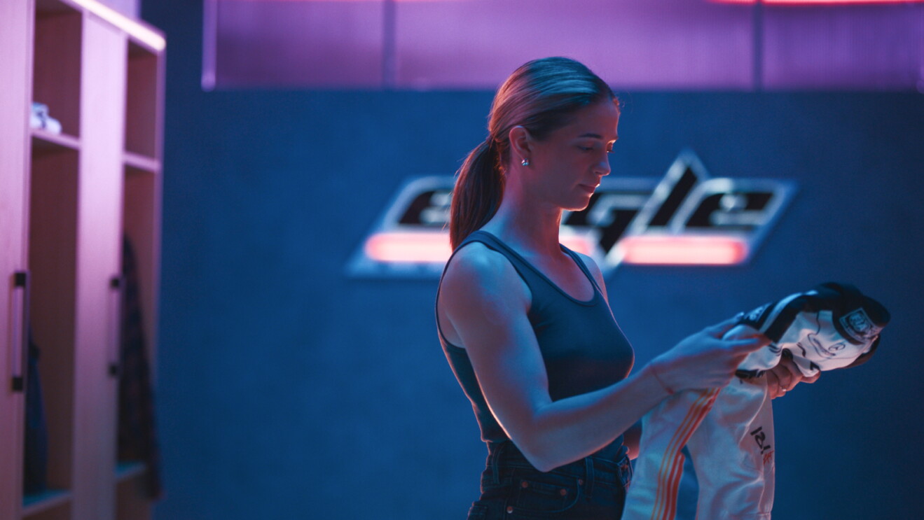 A young woman stands in a locker room bathed in pink and blue neon light. She looks down thoughtfully while holding a white sports jersey
