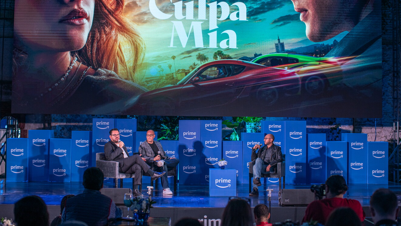 En un escenario con cajas con el logo de Prime, aparecn tres personas sentadas que no se pueden identificar y encima de ellas el cártel de la película Culpa Mía. 