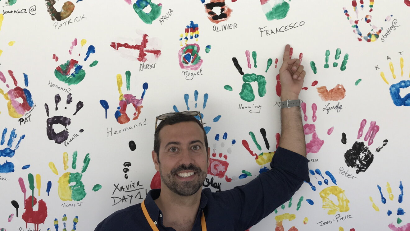 Ritratto a mezzo busto di Francesco Magini, responsabile del Kindle Store per l’Europa. L'uomo indossa gli occhiali da sole sulla testa e con il braccio sinistro tenuto in alto su di sè indica il muro alle sue spalle, ricoperto di impronte colorate di mani. 