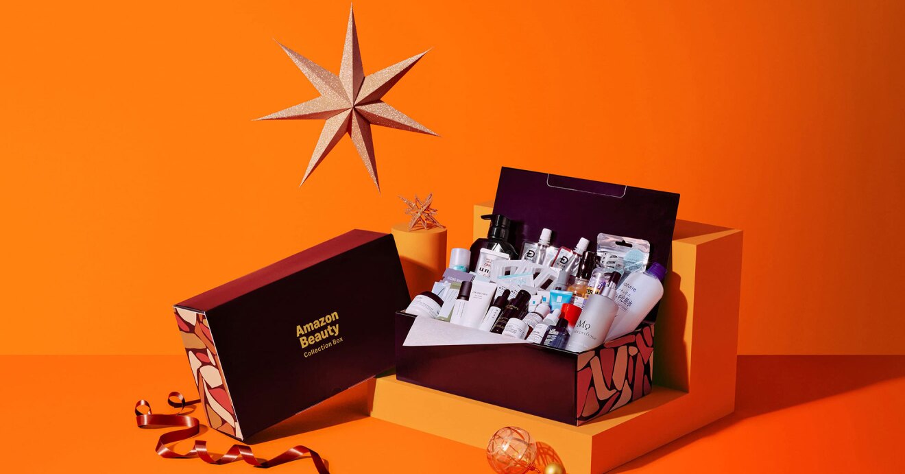 オレンジ色の背景に、星のオブジェとオレンジの台座に置かれたコスメのボックス。蓋が開いて中にたくさんの基礎化粧商品がつまっている。左側の外箱に「Amazon Beauty」の文字。