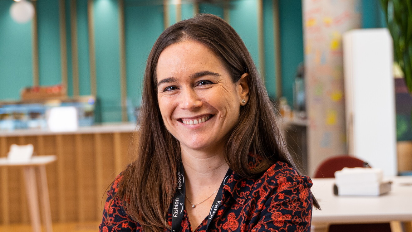 Lila Fernández es responsable de moda de los marketplace de Amazon en España e Italia.  De fondo está la cantina con mesas y los mostradores de comida. Lila mirando a cámara y sonriendo. Lleva un badge con una cadena de color negro. Una camisa azul con estampado de color rojo. Lleva pendientes y el pelo castaño, liso y largo. 