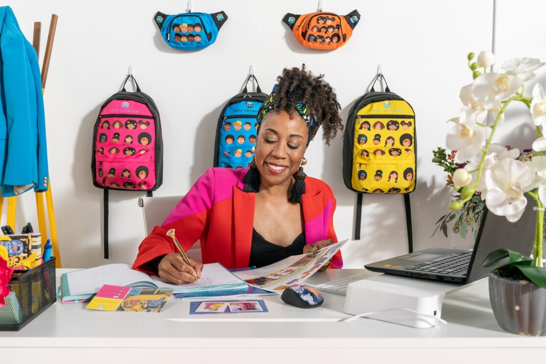 Cara sits at her desk and smiles as she writes in a notebook. EPIC Everyday product hangs on the wall behind her.
