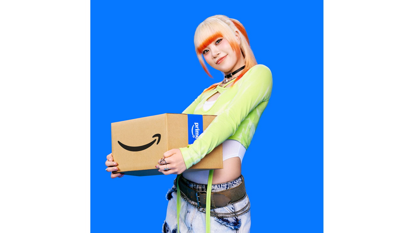 ハイトーンカラーヘアの若い女性が、Amazonロゴ入りダンボール箱を両手で抱えて身体の前で持っている。