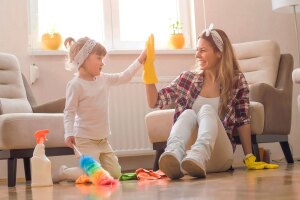 A mom smiles and gives her daughter a high five as they sit on the floor next to cleaning supplies in a living room.