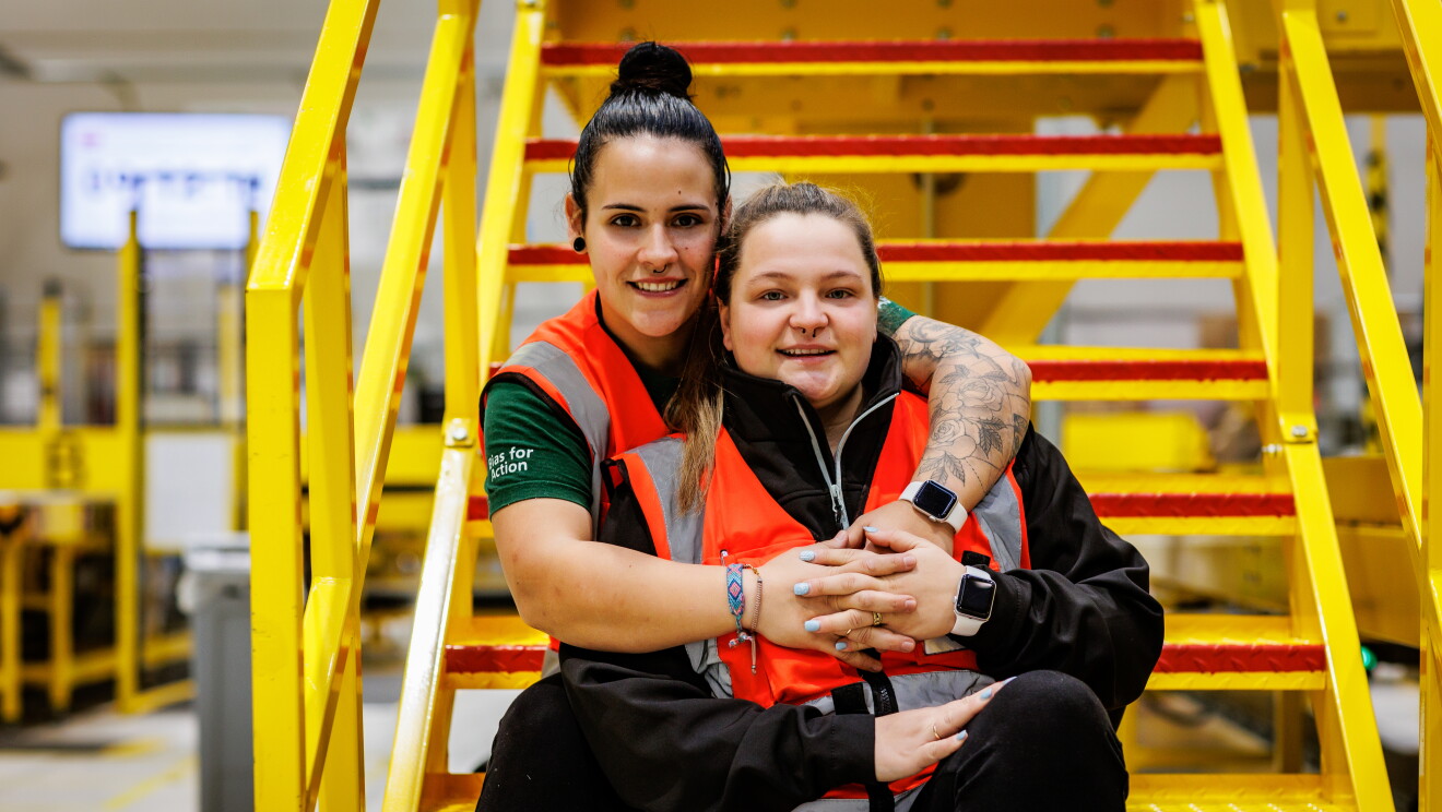 La pareja de la foto se llaman igual, Cristina. Están dentro de un centro logístico, sentadas en una escalera de color amarillo. Ellas dos están en los peldaños. una de ellas le agarra por la espalda y la de delante le agarra la mano. Llevan el pelo recogido con una coleta y con un moño alto, los zapatos de seguridad, pantalón negro y chaleco de seguridad de color naranja. Las dos tienen un piercing en la nariz. 