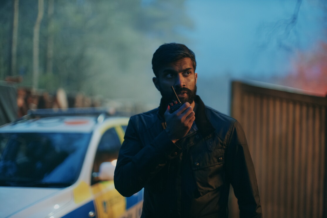 A bearded police officer holding a walkie talkie and standing next to a police car