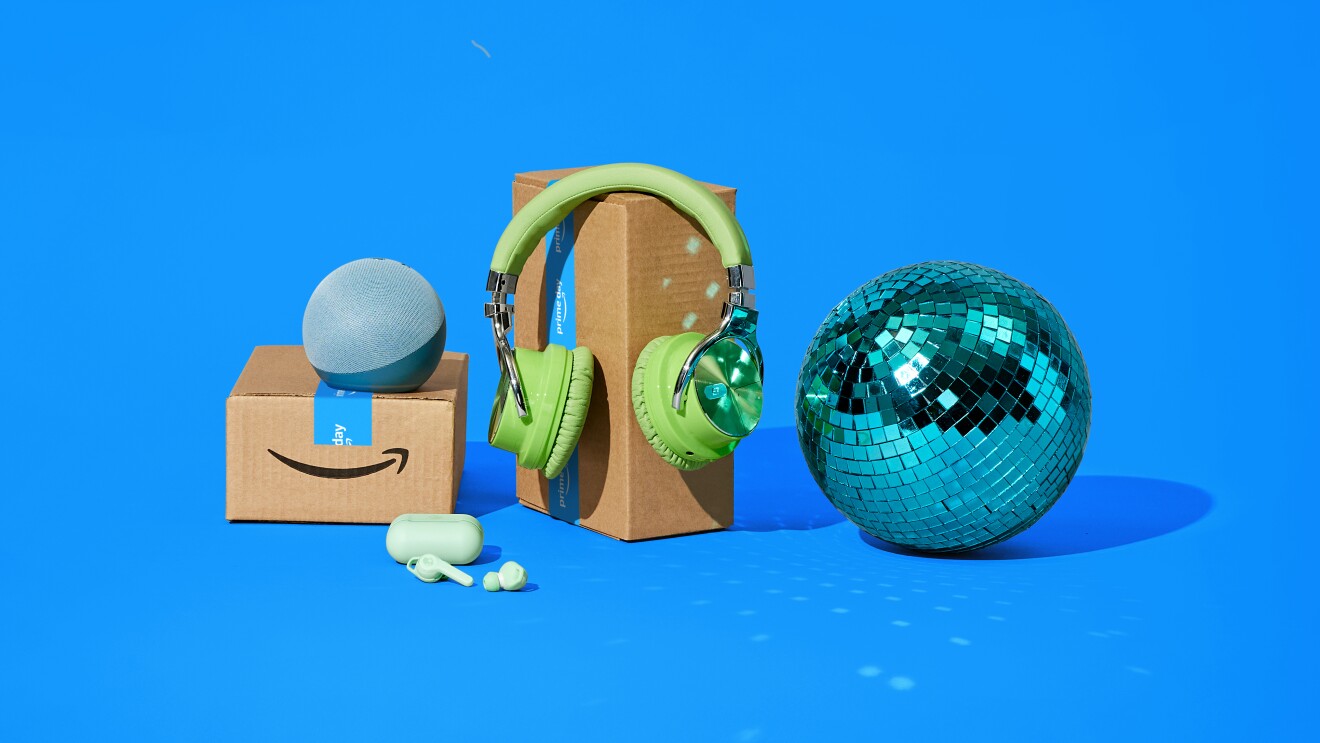 Fotomontaje, sobre un fondo azul, dos cajas de Aamzon, una con dos dispositivos Amazon apoyados, la caja grande con unos auriculares verdes y al lado derecho una bola de fiestas de discoteca.