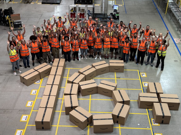 Mitarbeitende stehen im Logistikzentrum um die aus Paketen gelegte Zahl 16+