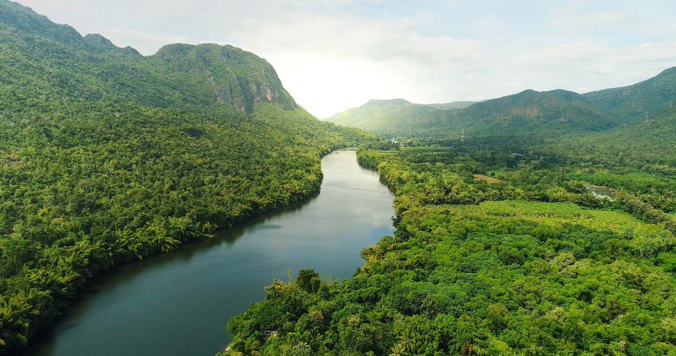 Foto de un paisaje. Se ve un río con agua verde rodeado de un bosque frondoso de árboles. Se ven cuatro montañas y en el fondo el sol.