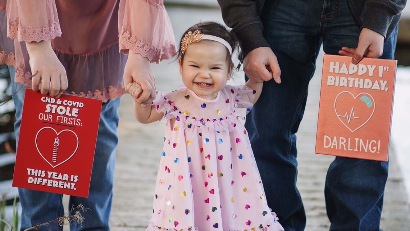 A toddler stands holding her mom and dad's hands. Her parents hold large greeting cards on either side of her. 