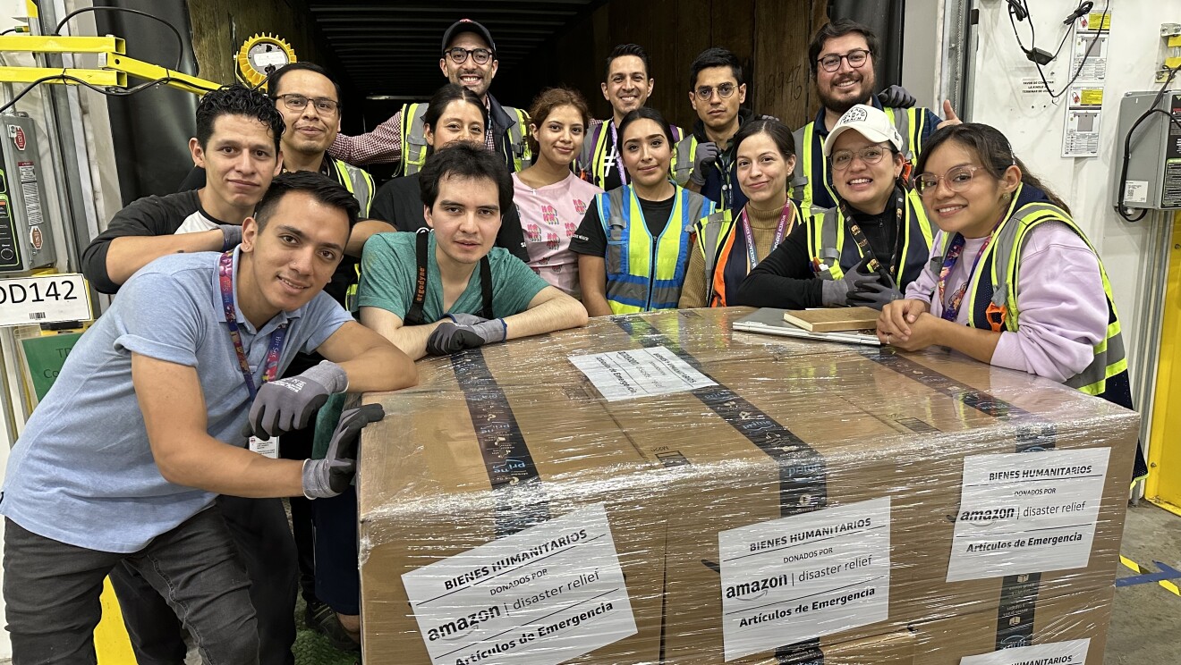 Trabajadores de Amazon delante de un palé con cajas de Amazon etiquetadas con un cartel que pone: Bienes humanitarios, artículos de emergencia.