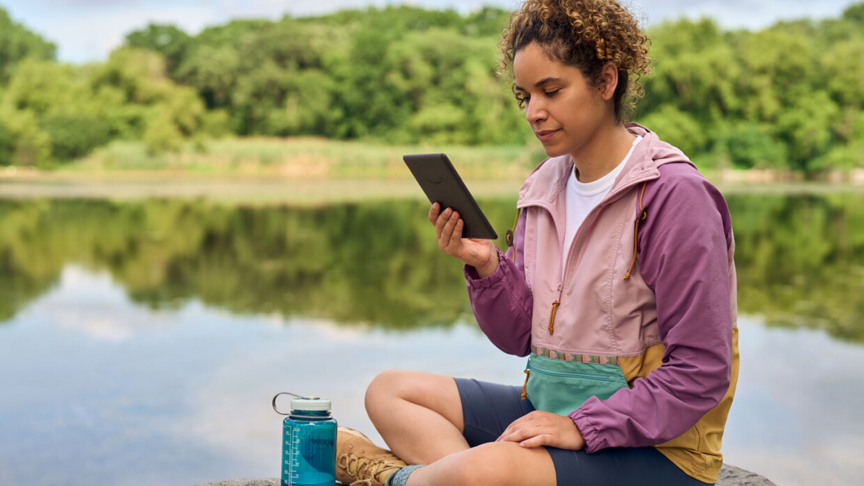 A woman sat cross-legged by a lake holding a Kindle.