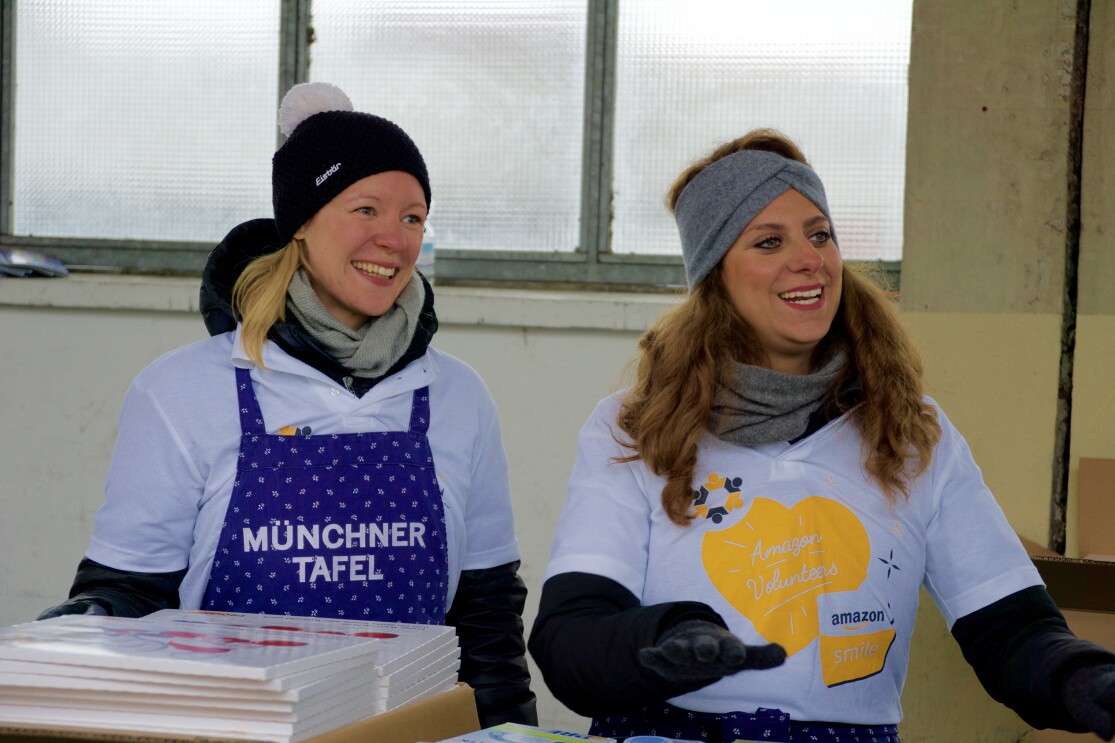 Zwei Junge Frauen geben an der Großmarkthalle Weihnachtskalender aus. Eine trägt eine Schürze mit Aufschrift "Münchner Tafel", die andere ein T-Shirt mit Aufdruck "Amazon Volunteesrs"