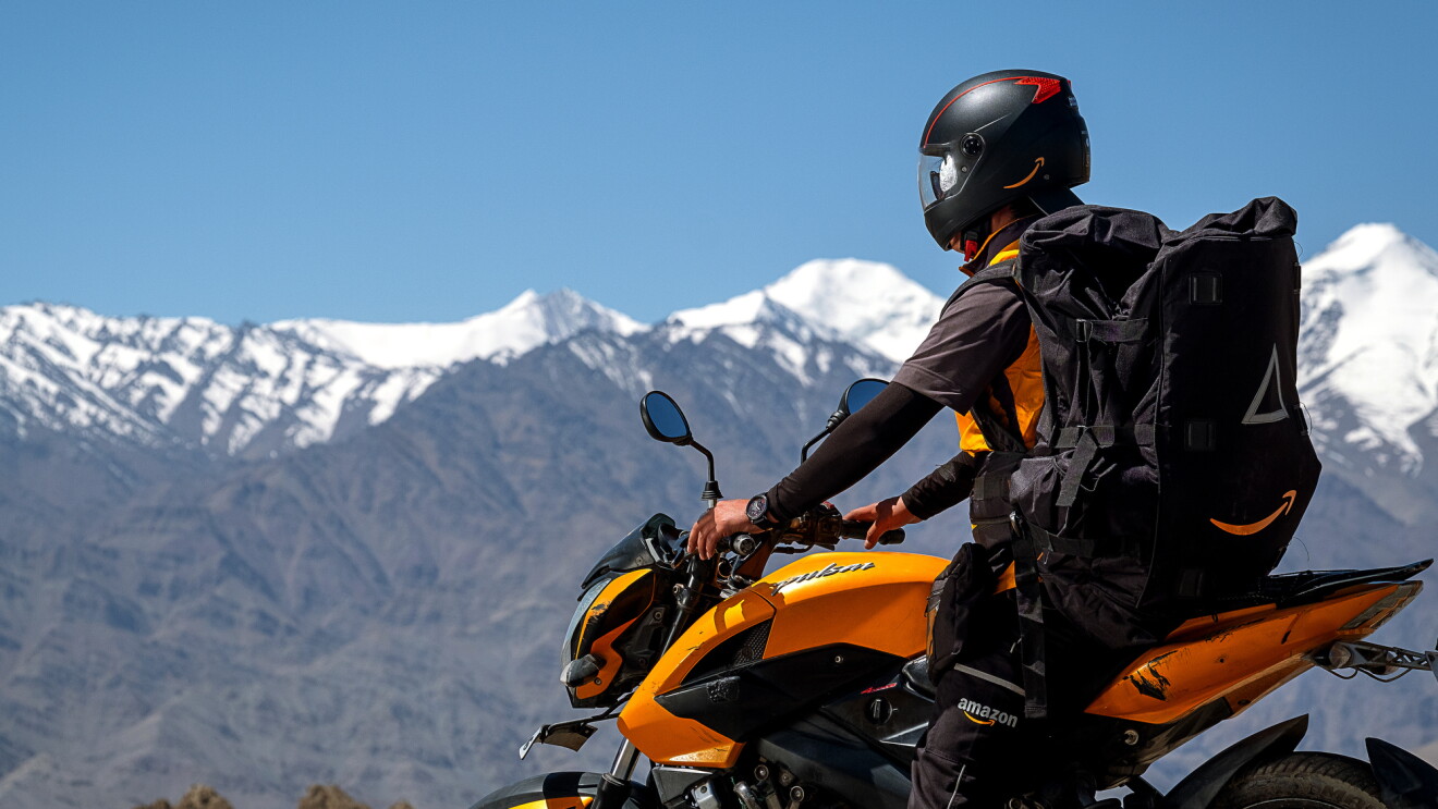A helmeted motorcyclist wears a large black backpack with the Amazon smile logo. Snow-capped mountain peaks are in the background of the image.