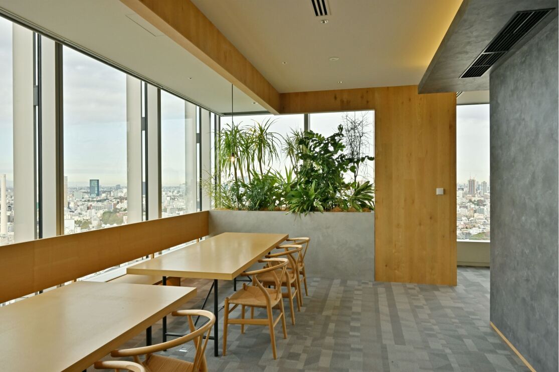 L字型のスペースの左側にテーブルが２つ置かれている。その横に大きな窓と植物が植えられたスペースがある