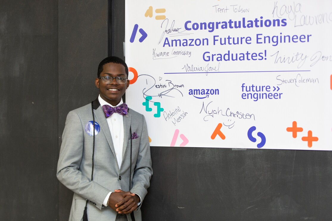 A student wears a suit and bow tie, and smiles, in front of an Amazon Future Engineer poster at his school.