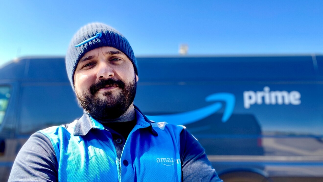 Nick Gallina wears his blue Amazon attire while smiling in front of his delivery van that features the Amazon logo.