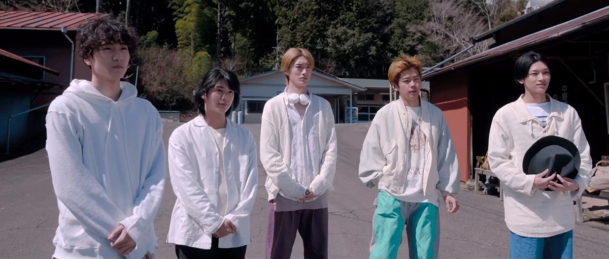 木々が茂る山、奥の方の平屋や納屋を背景に、前庭らしき敷地内に建つ5人の若者たち。デザインの異なる白い長袖の上衣を着用し、水色や紺、黒など異なる色のパンツを履いている。