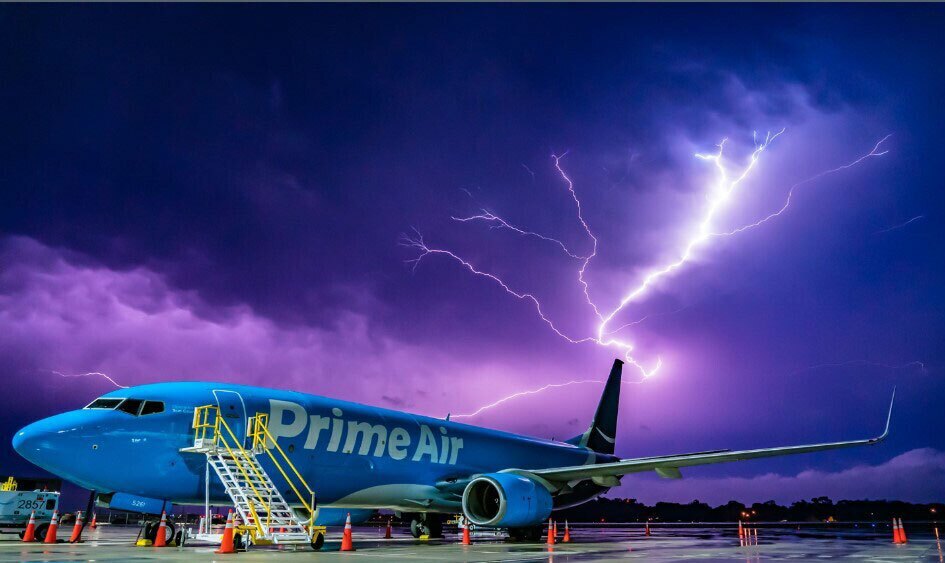 An image of a Prime Air plane being loaded on the runway. The sky is dark and purple behind the plane with a large lightening strike moving through the clouds.