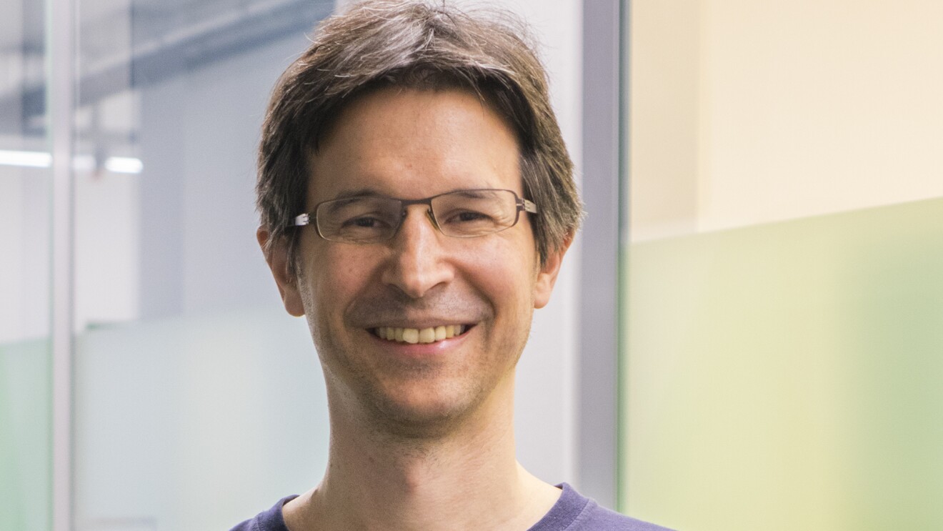 Ein Mann mit braunen Haaren, Brille und einem blauen T-shirt lacht in die Kamera.