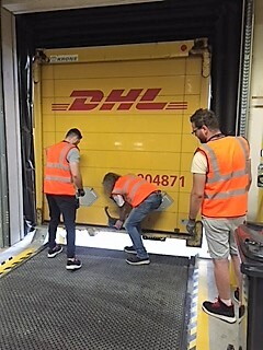 3 Versandmitarbeiter mit orangenen Warnweste sind vor einer Laderampe des Logistikzentrum zu sehen. Die Mitarbeiterin in der Mitte schließt gerade die Versandklappe des Lkws.