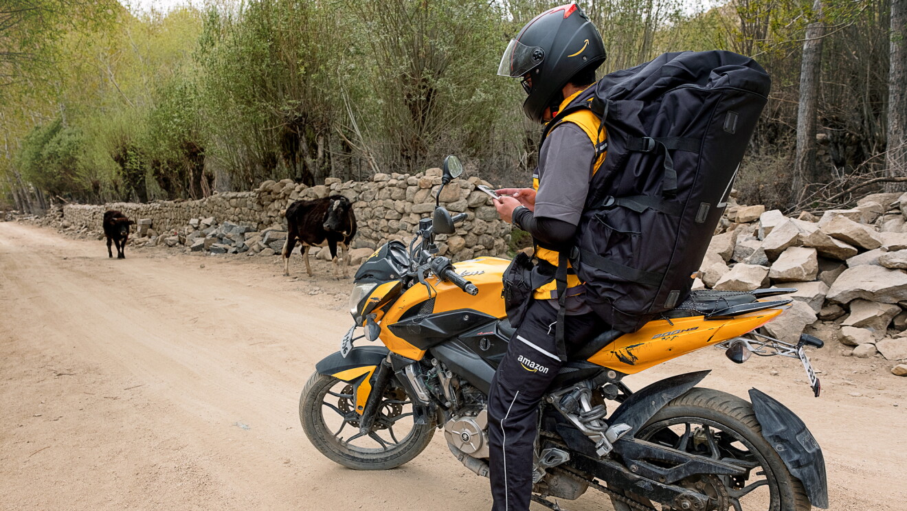 A motorcyclist wearing a large black backpack and an Amazon delivery uniform stands astride a motorcycle on a dirt road. He looks down and checks his smartphone. Two cows are in the background of the image.