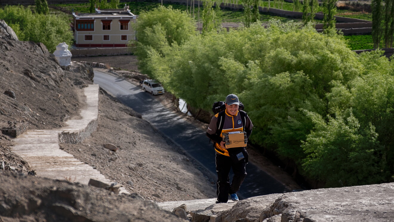 A smiling man in an Amazon delivery uniform carries a package up a staircase of at least 100 stairs that snakes along a hillside up from a road below. Trees and buildings are in the background of the image.