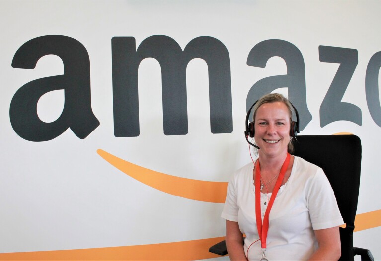 Eine Amazon Kundenservice-Mitarbeiterin mit Kopfhörer vor einer Wand mit Amazon Schriftzug.