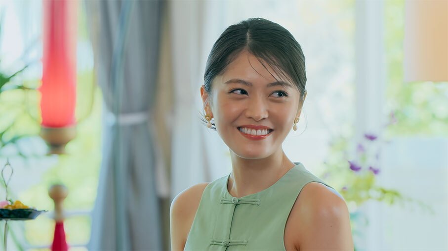 光が入る明るい屋内で微笑む女性。ノースリーブの民族衣装を着用。背後にカーテンや植物が見える。