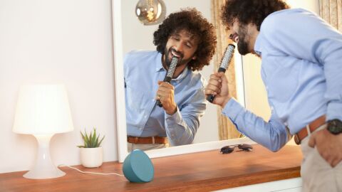 Man with curly hair sings passionately into hairbrush microphone in front of mirror