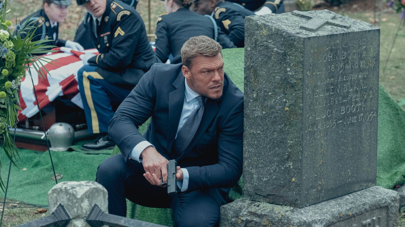 jack reacher crouched behind a tombstone holding a gun in the prime video series reacher