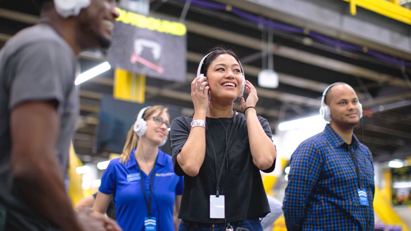 Four people wearing headphones and smiling during an Amazon fulfillment center tour. 