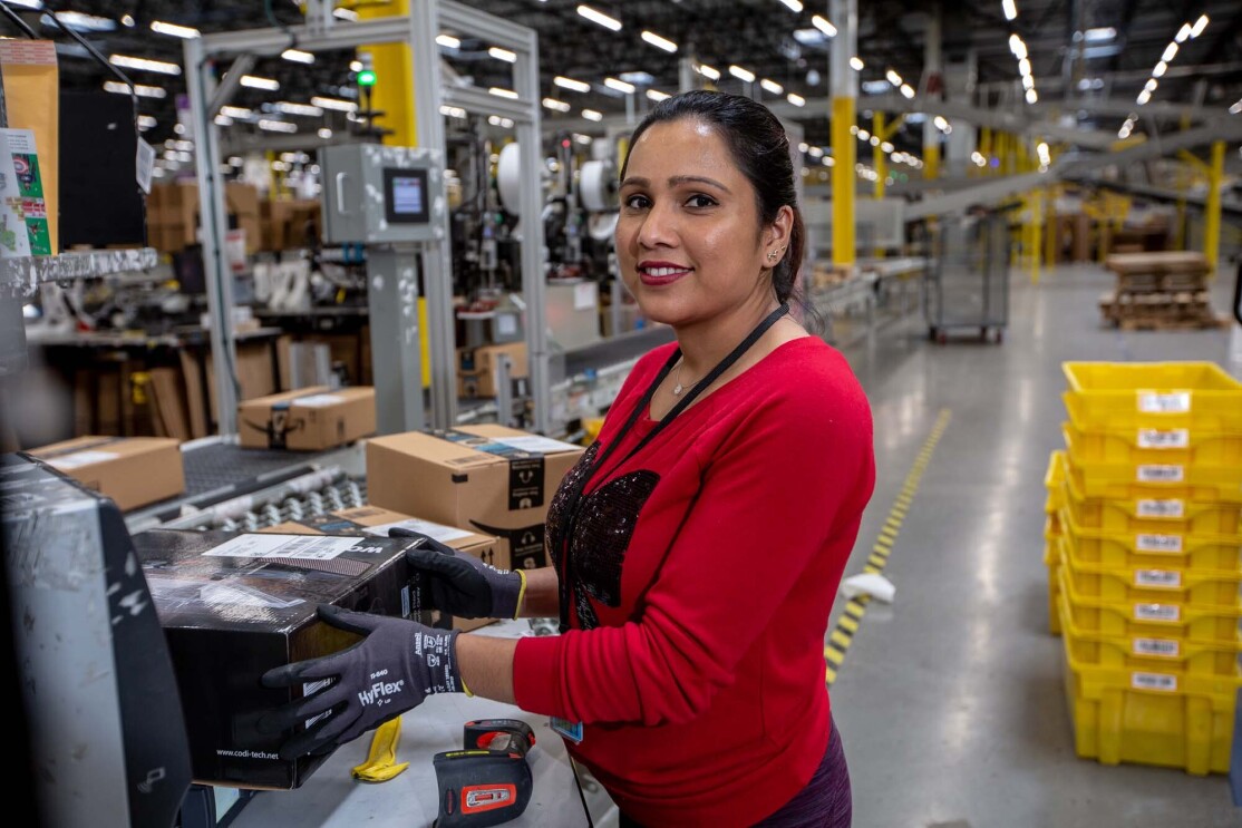 An Amazon associate holds a product near a conveyor belt. Tools sit on the countertop near her. She wears a red long-sleeve tee shirt and work gloves as she works at a Columbus, Ohio fulfillment center, CMH1