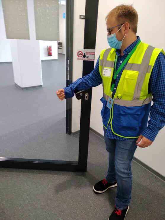 Mitarbeiter in Sicherheitsweste und Mundschutz öffnet die Türe mit der Armbeuge.