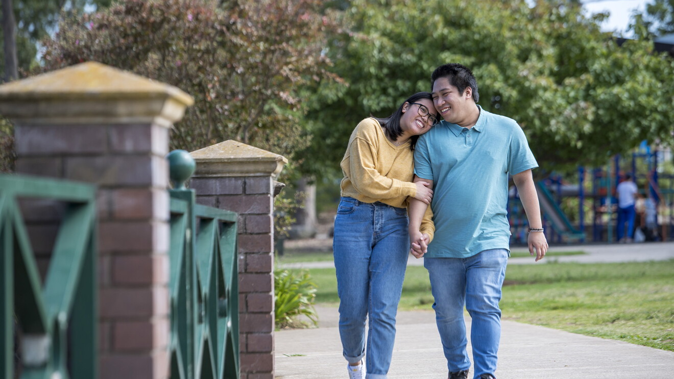 An image of a couple walking down the sidewalk together, holding hands and smiling. The woman is resting her head on the man's shoulder. 