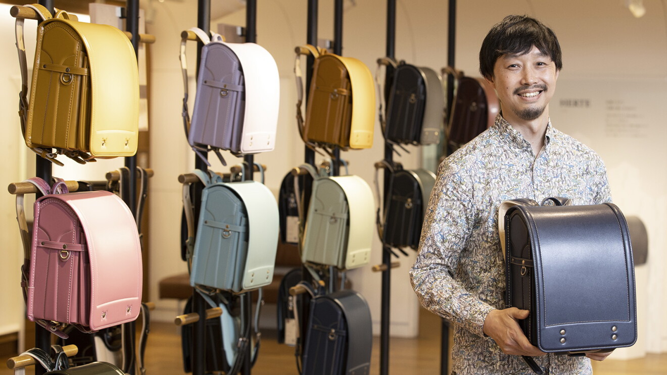 カラフルなランドセルが展示されている前に立つ口髭のある男性。紺色のランドセルを手に持っている