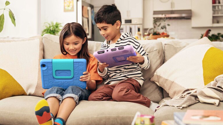 An image of two kids playing with their tablets sitting on a sofa.