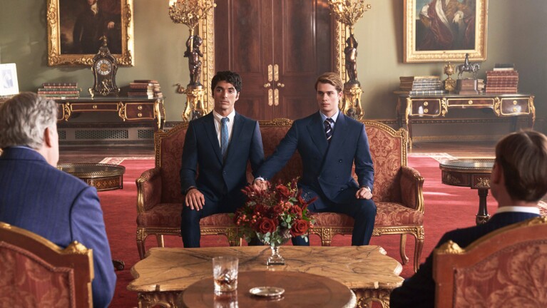 A scene of Red White and Royal Blue showing two men sitting side to side in a living room wearing suits and holding hands. 