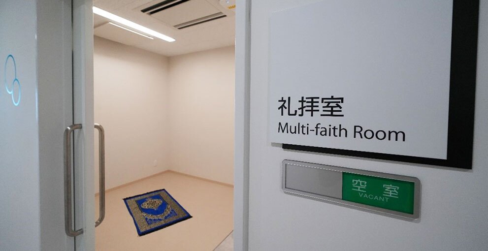 開いた白いドアの奥の小部屋に、青地に金色模様の長方形のじゅうたんがある。ドアの右側の壁には「礼拝室 Multi-faith Room」の表示。その下に銀のプレートがあり、緑色で「空室」の表示。