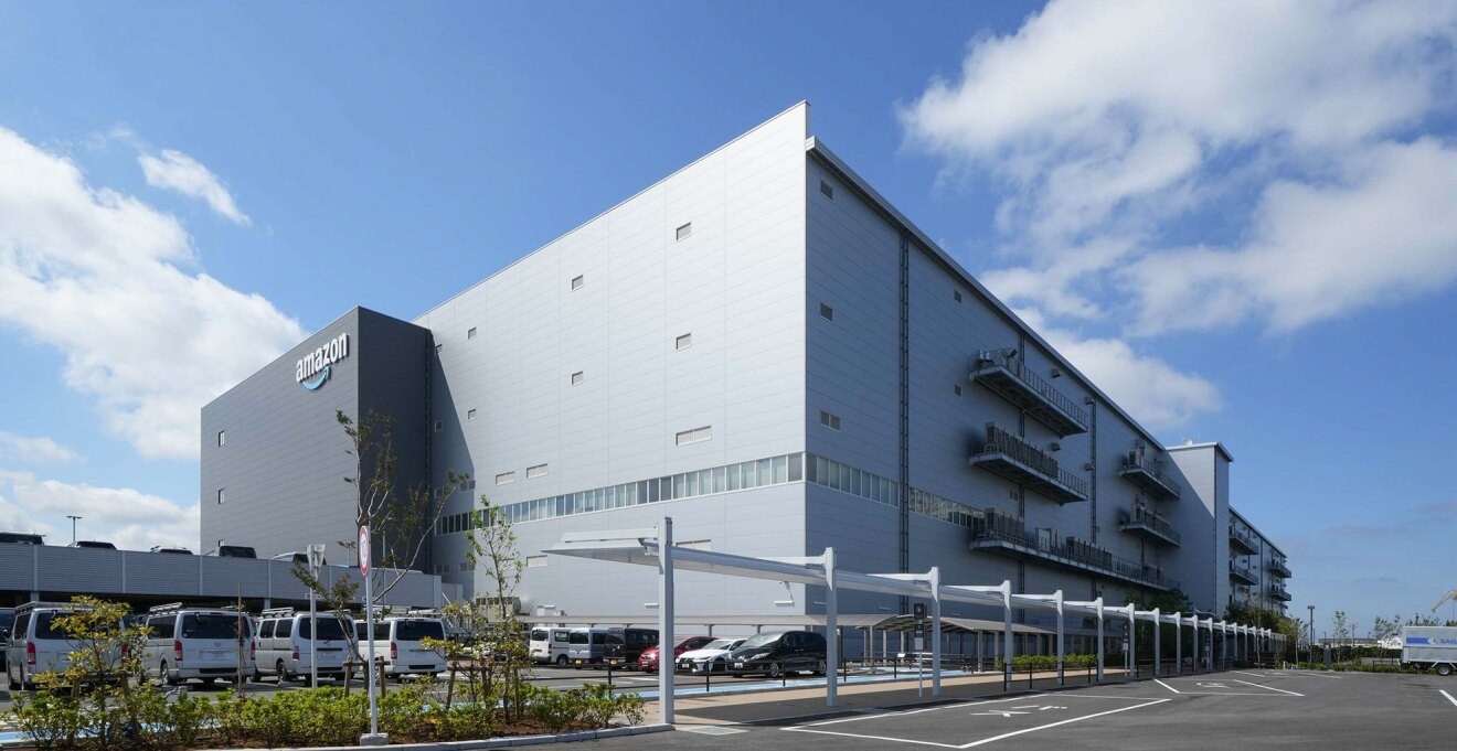 青い空をバックに建つ大きな建物。左側のダークグレーの壁にAmazonロゴ。右側の壁は薄いグレー。建物の手前に駐車場とバス乗り場の屋根。
