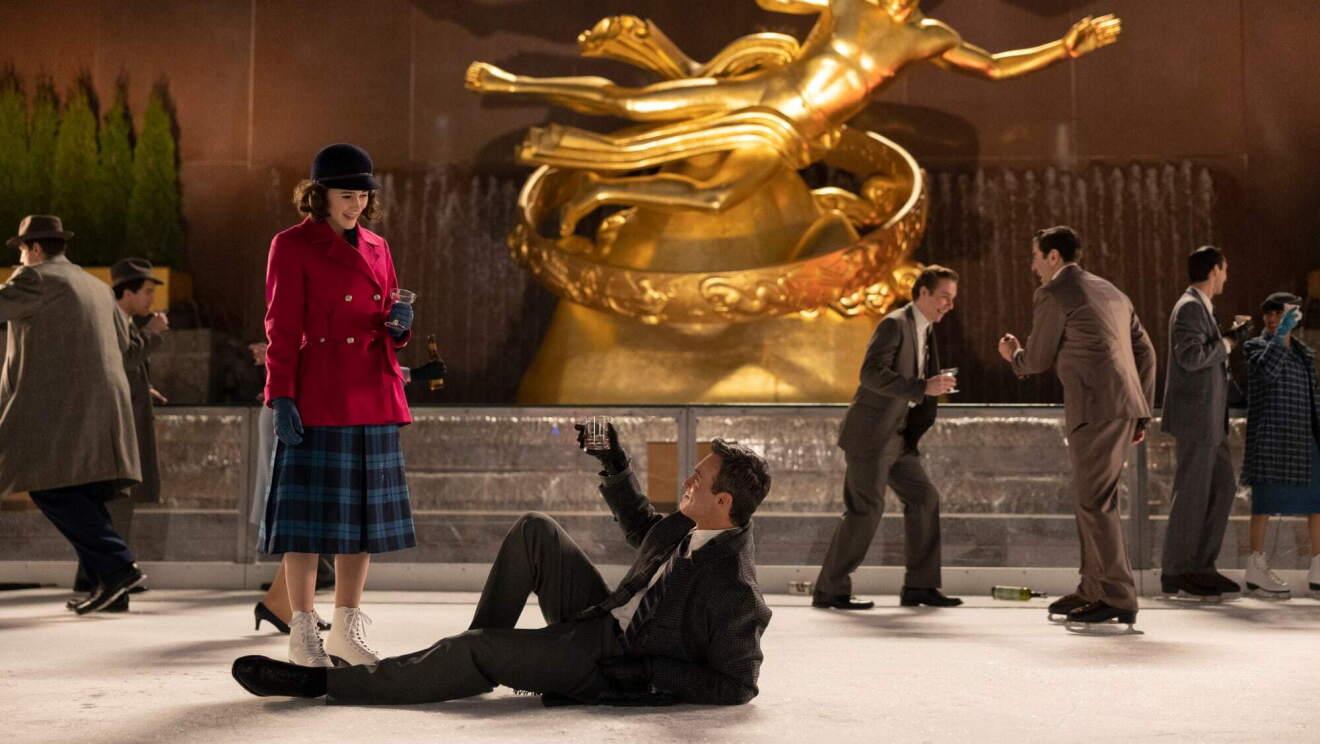 An image of Midge Maisel standing on an ice rink with a man on the ground below her reaching for her hand.